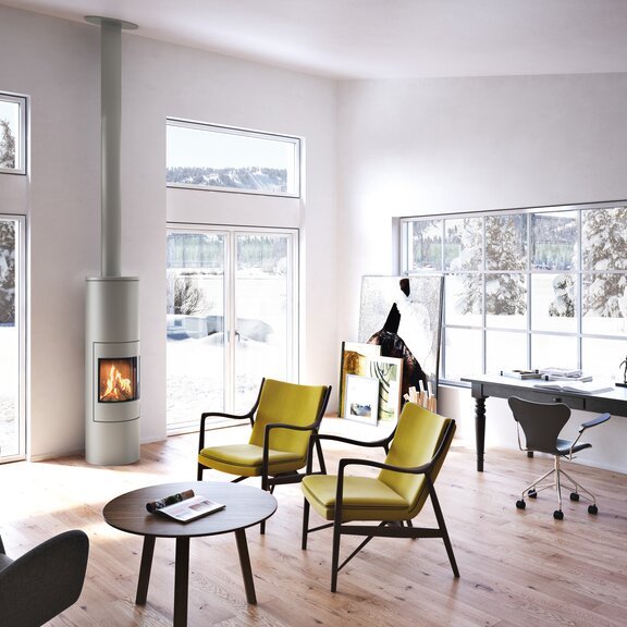 Poêle à gaz VIVA 160 L en nickel avec porte en acier dans un bureau avec de grandes fenêtres entourées d'un paysage hivernal enneigé