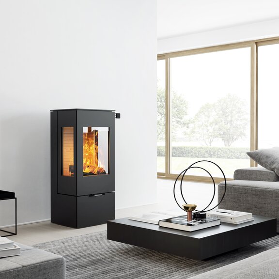Poêle à bois NEXO 100 en noir avec porte en acier et deux vitres latérales dans un salon moderne et lumineux