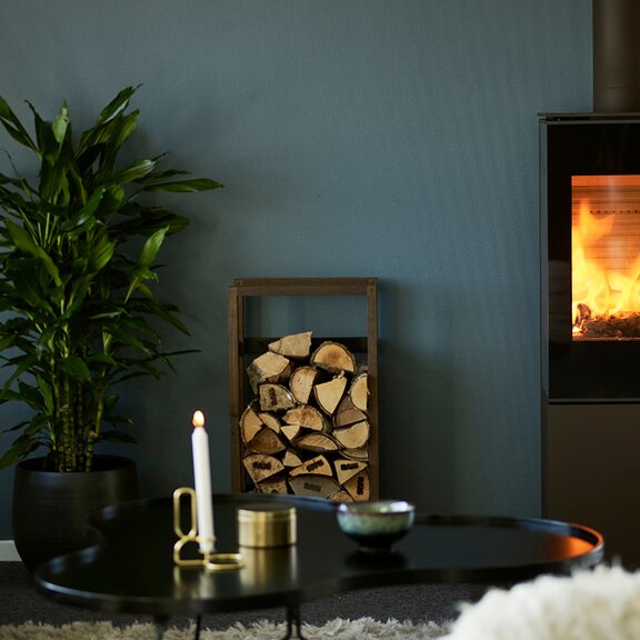 Poêle à bois NEXO 120 en noir avec porte de foyer plein verre et feu tranquille devant un mur couleur pétrole dans un appartement de style
